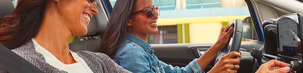 Two women, one driving and one passenger, smiling while traveling in a car.