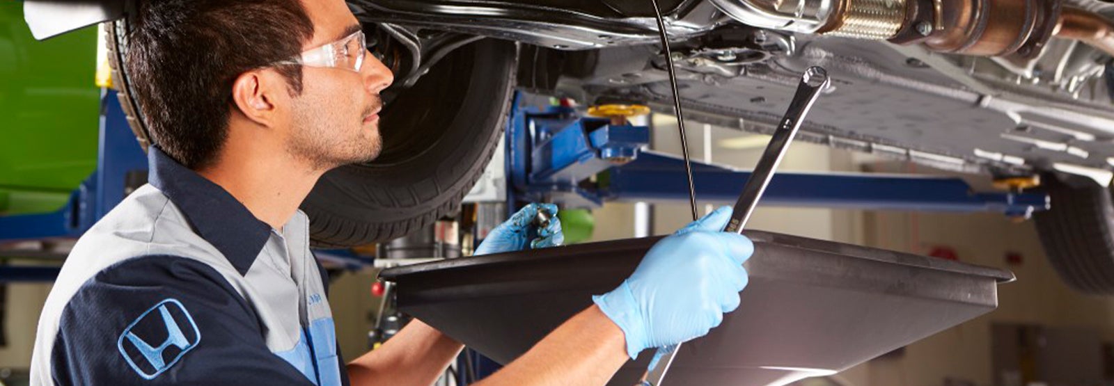 A mechanic in safety glasses and a Honda uniform changes oil on a lifted car, holding a wrench and drain pan.