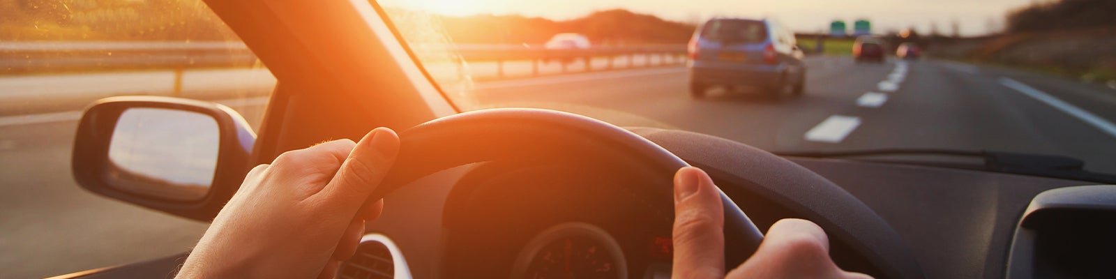 Driver's hands on a steering wheel, driving on a highway at sunset with other cars visible.