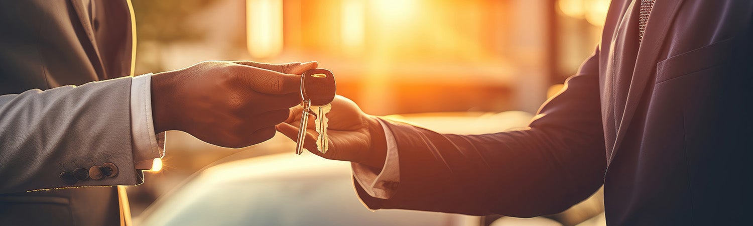Two people in suits exchanging car keys, backlit by a bright golden sunset.